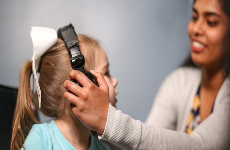 A young girl has headphones placed on her head by a speech-language pathologist.